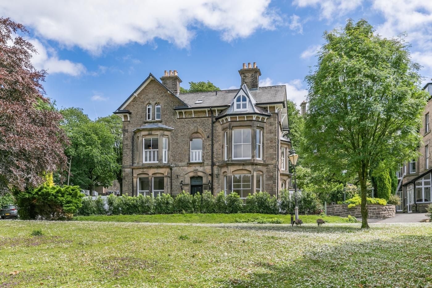 A large, historic stone building with multiple windows and a prominent bay window, surrounded by well-maintained gardens and trees.