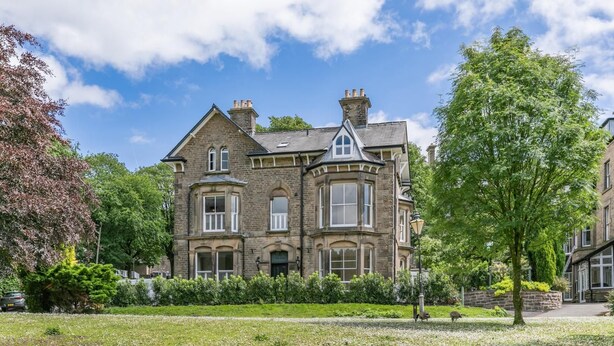 A large, historic stone building with multiple windows and a prominent bay window, surrounded by well-maintained gardens and trees. - 3 Bed in Buxton (oc-p32528) (Buxton)
