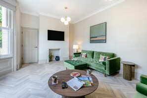 A living room with a green sofa, a round coffee table with newspapers and remote controls, a fireplace, and a flat-screen TV mounted on the wall. - 3 Bed in Buxton (oc-p32528) (Buxton)