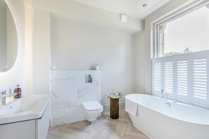 Une salle de bain moderne avec un mur en marbre, des toilettes, un lavabo, une baignoire et une fenêtre avec des stores.