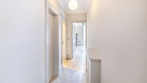 Un couloir moderne aux murs blancs, doté d’un parquet en chevrons et d’un luminaire suspendu. Des portes donnent accès aux autres pièces.