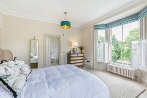 Une chambre à coucher avec un grand lit, une commode, un miroir, une fenêtre avec des volets et un radiateur.