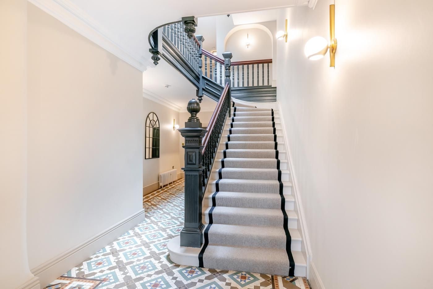 A grand staircase with a patterned carpet, a black balustrade, and a white wall with a light fixture.