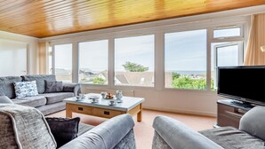 A living room with a sofa, coffee table, and TV, large windows with a view, and a wooden ceiling. - 4 Bed in Padstow (oc-p00401) (Trevone)