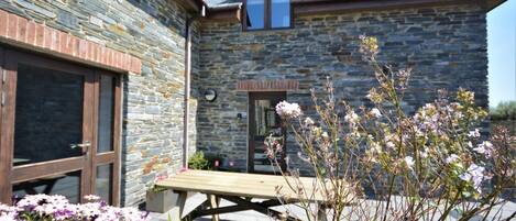 A stone building with a wooden door and window, a wooden bench, and flowering plants in the foreground.