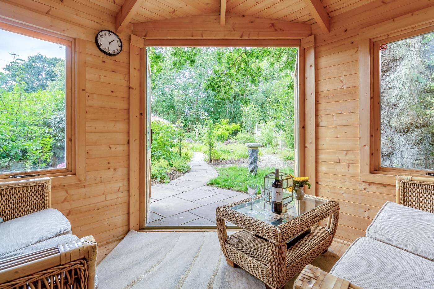 Un intérieur en bois avec une porte vitrée donnant sur un sentier de jardin, des meubles en osier et une table avec une bouteille et des verres.
