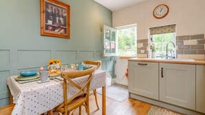 A kitchen with a dining table set for two, a clock on the wall, and a window with a view of greenery.