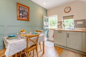 A kitchen with a dining table set for two, a clock on the wall, and a window with a view of greenery.