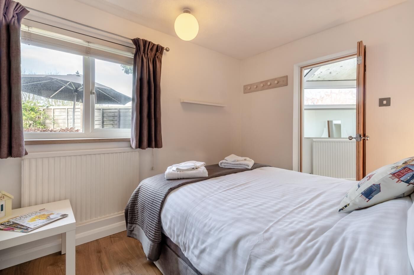Une chambre à coucher avec un lit, une table de chevet sur laquelle se trouve un magazine, une fenêtre avec des rideaux et une porte.