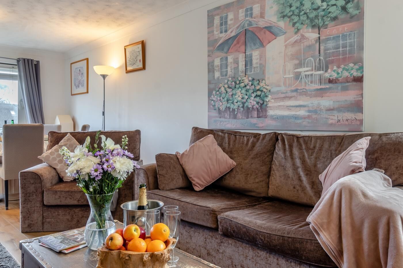 A cozy living room with a brown sofa, a coffee table with fruits and a vase of flowers, a lamp, and a painting of a cafe scene.