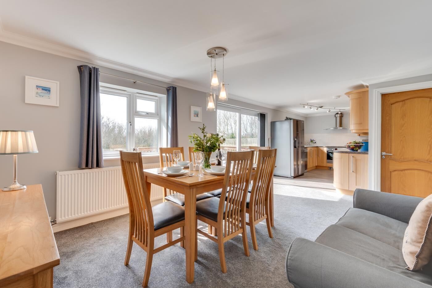 A modern living room with a dining table set for four, a sofa, a kitchen in the background, and large windows.