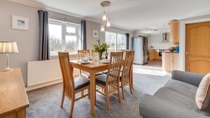 Una sala moderna con una mesa de comedor preparada para cuatro personas, un sofá, una cocina al fondo y ventanas amplias.