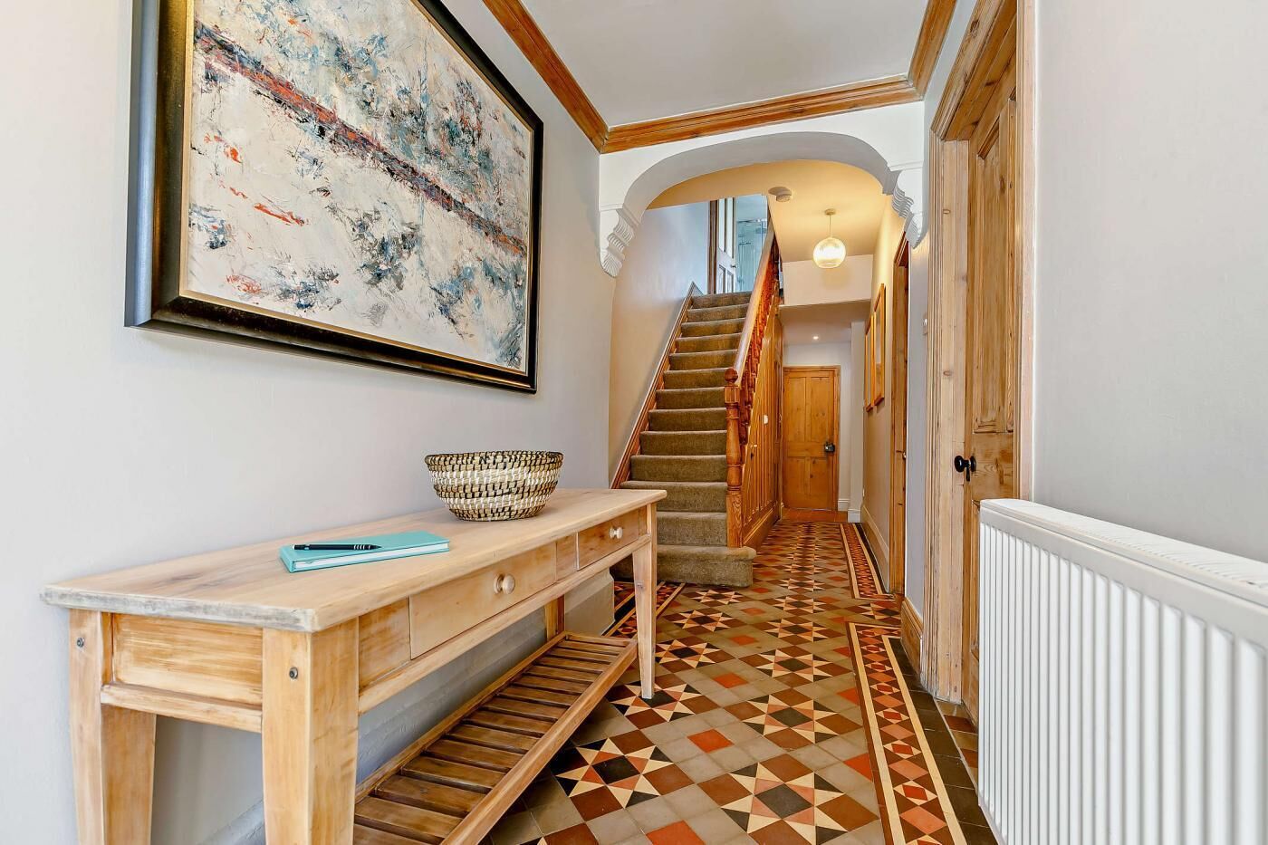 A hallway with a patterned floor, a wooden console table, a large abstract painting, and a staircase leading to another room.