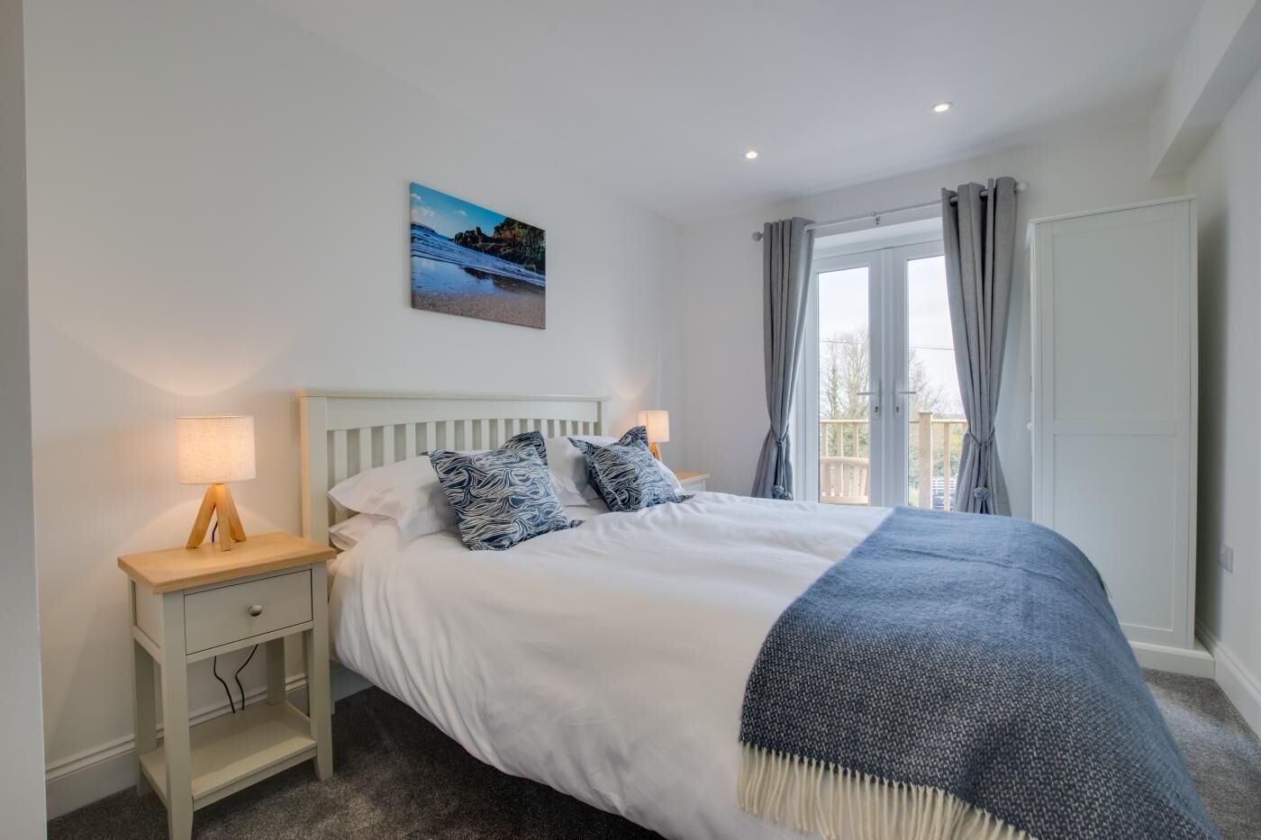 A bedroom with a bed, bedside tables, a nightstand, a window with curtains, and a view of the outdoors.