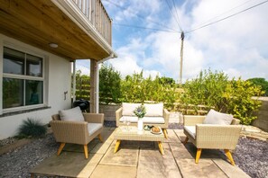 An outdoor patio with wicker furniture, a barbecue grill, and a view of a garden with trees and a fence. - 3 Bed in Little Petherick (oc-p00899) (Little Petherick)