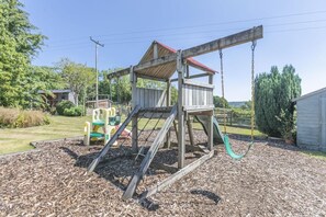 Een houten schommelset met een rood dak, een glijbaan en een klimconstructie in een achtertuin met mulch als grondbedekking.