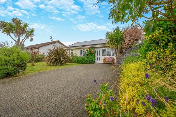 A residential house with a paved driveway, surrounded by lush greenery and flowering plants. - 3 Bed in St. Merryn (oc-p00114) (Saint Merryn)