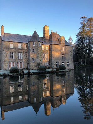 Un bâtiment historique en pierre, doté de nombreuses fenêtres, dont le reflet se mire dans l’eau.