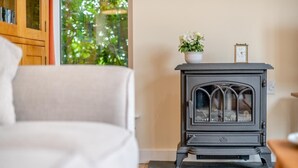 Ett mysigt vardagsrum med en grå vedkamin, en vit soffa och ett fönster med utsikt över grönskan.