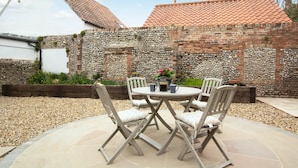 Une terrasse extérieure avec une table ronde et quatre chaises, entourée d’un mur en pierre et d’un espace jardin.