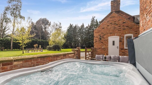 A hot tub in a backyard with a brick building, a wooden deck, and a picnic table. - 4 Bed in Bronington (oc-g32599) (Bronington)