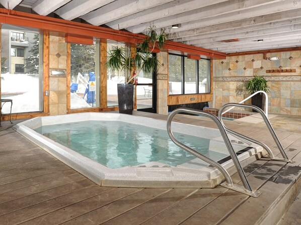 An indoor swimming pool with a wooden deck, large windows, and a stone wall.