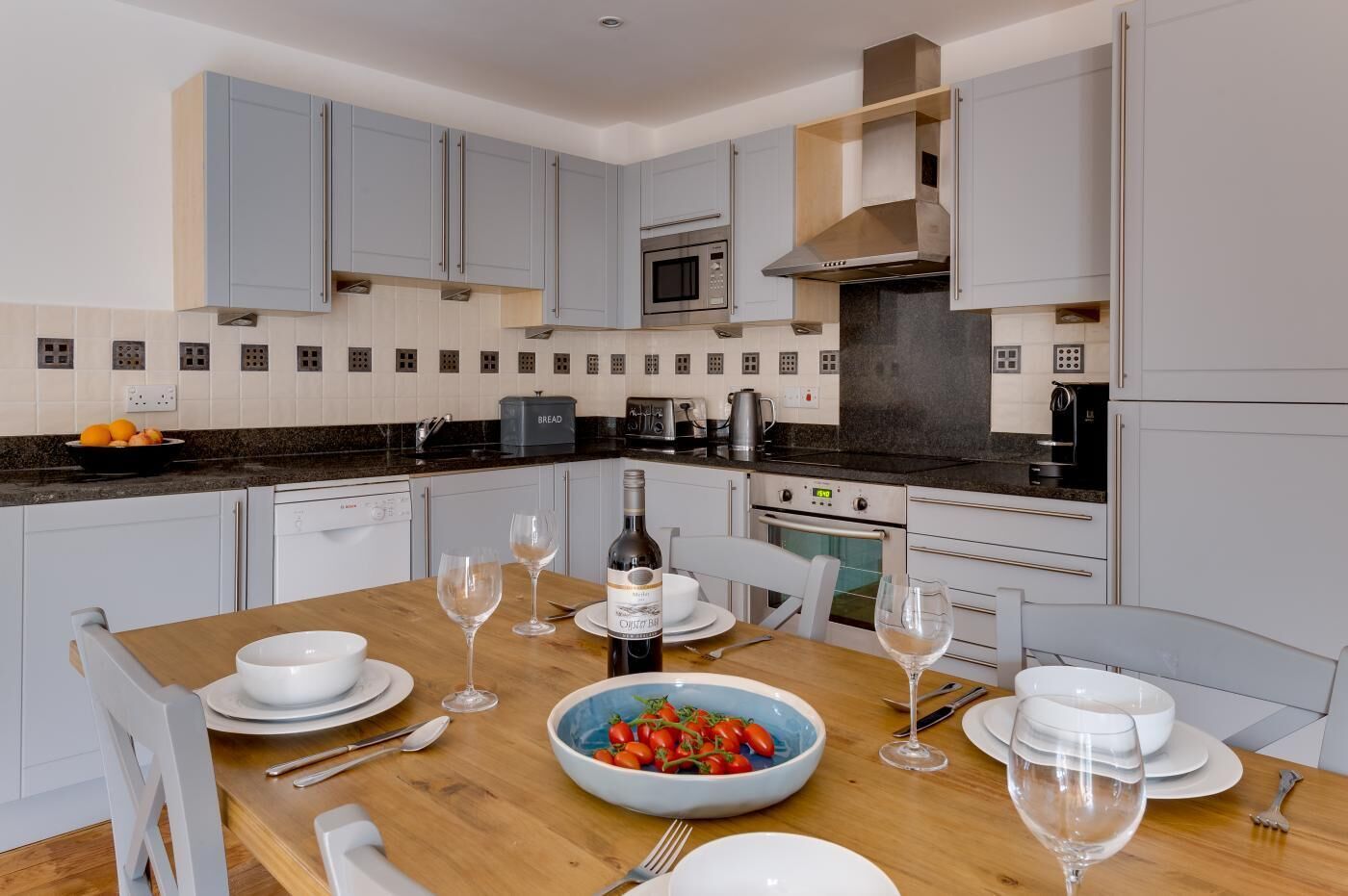 A modern kitchen with grey cabinets, a wooden dining table set with plates, glasses, and a bowl of tomatoes.