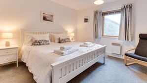 Een slaapkamer met een wit bed, twee nachtkastjes, een stoel, een raam met gordijnen en een radiator.