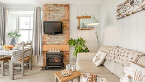 A cozy living room with a stone fireplace, a television mounted above it, a sofa with patterned cushions, and a dining area with a table set for breakfast. - 1 Bed in Tintagel (oc-pc084) (Tintagel)