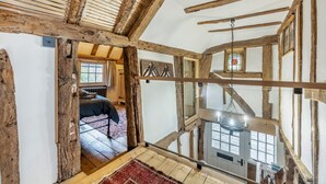 A rustic room with exposed wooden beams, a bed with a red and blue patterned rug, and a view of a lower room through a window. - 2 Bed in Battle (oc-f32643) (Battle)