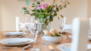 Mesa de comedor dispuesta con platos, cubiertos, vasos y un centro de mesa con flores y pan.
