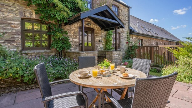 A stone building with a wooden table and chairs, surrounded by greenery and a wooden fence. - 2 Bed in Cantref (oc-t32629) (Cantref)