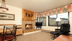 A living room with a fireplace, a television, a dining area with chairs, and a view of buildings through large windows. - Spacious Studio on Keystone Lake with Full Kitchen (Keystone)