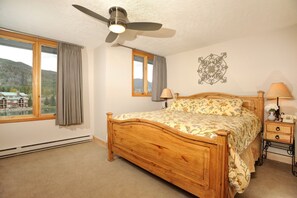 A bedroom with a wooden bed, a ceiling fan, a nightstand with a lamp, and a view of a house through a window. - Incredible and Spacious 2 Bedroom + Loft (Keystone)