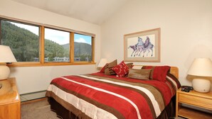 Une chambre avec un grand lit, une fenêtre donnant sur les montagnes, un tableau représentant des chevaux et une table de chevet avec une lampe.
