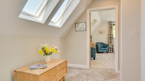 A room with a sloped ceiling, a wooden dresser, a vase of flowers, and a view into another room with a blue chair and curtains. - 2 Bed in Hemblington (oc-2134) (Hemblington)