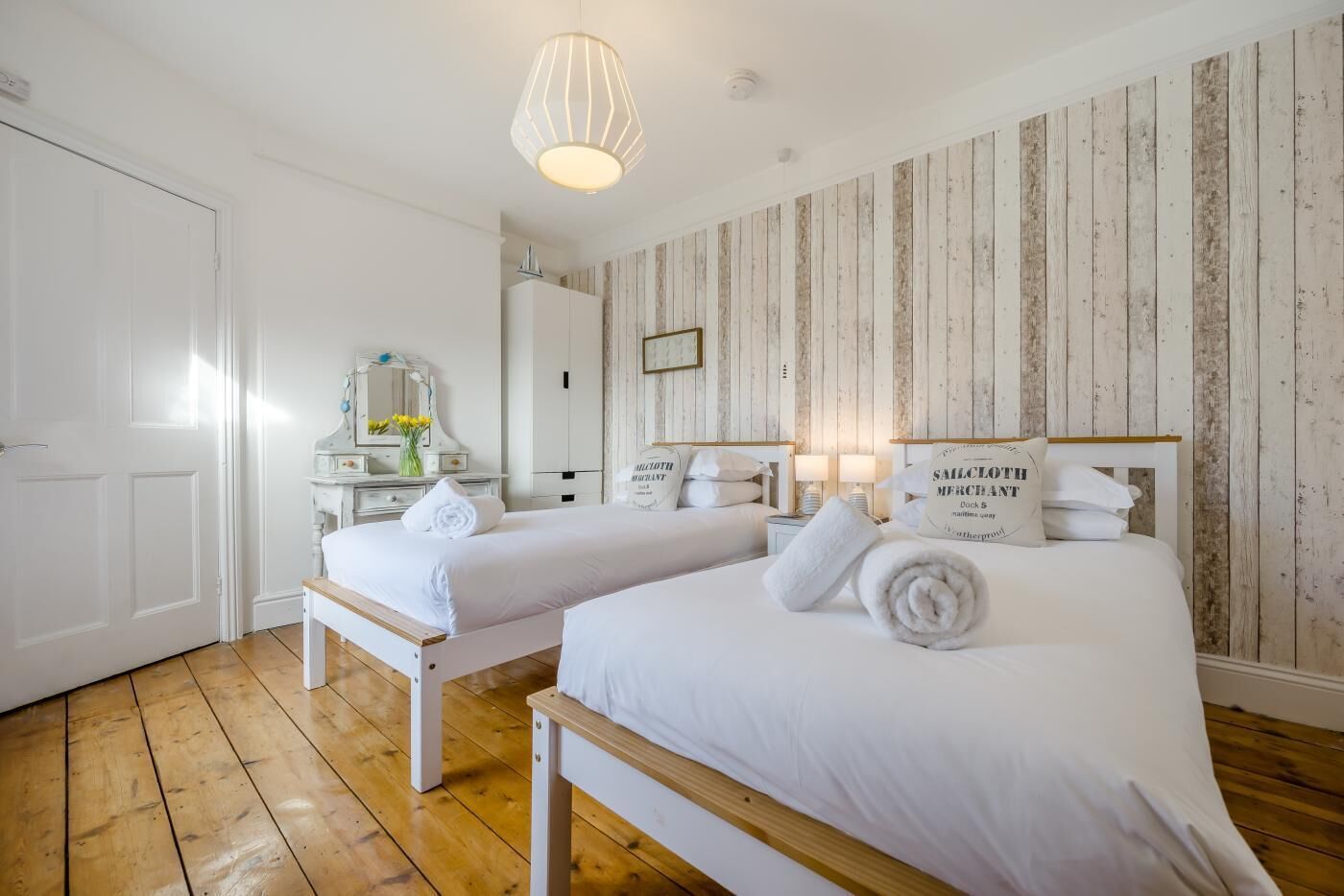 Une chambre d’hôtel avec deux lits, un plancher en bois, des murs blancs, un mur lambrissé, un miroir, une commode et un luminaire suspendu.