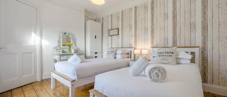 Ett hotellrum med två sängar, trägolv, vita väggar, en väggpanel av trä, en spegel, en byrå och en hängande lampa.