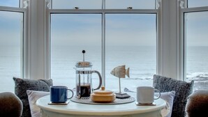 Et bord med kaffemaskine, kop og en dekorativ fisk, med udsigt til havet gennem store vinduer.
