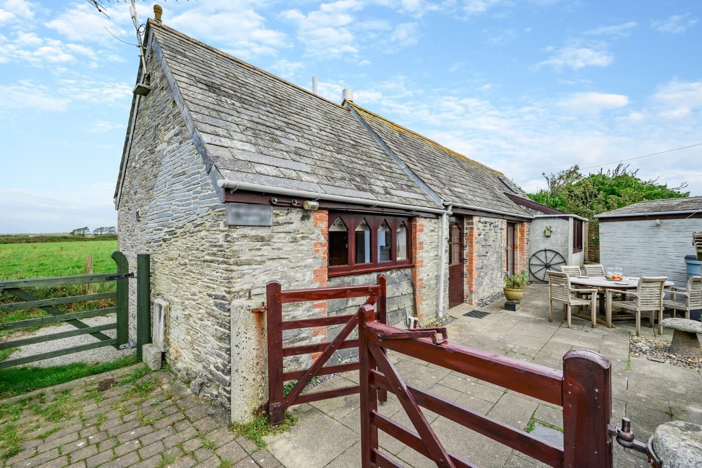 A stone farmhouse with a wooden gate, a wheelbarrow, and a table with chairs outside.