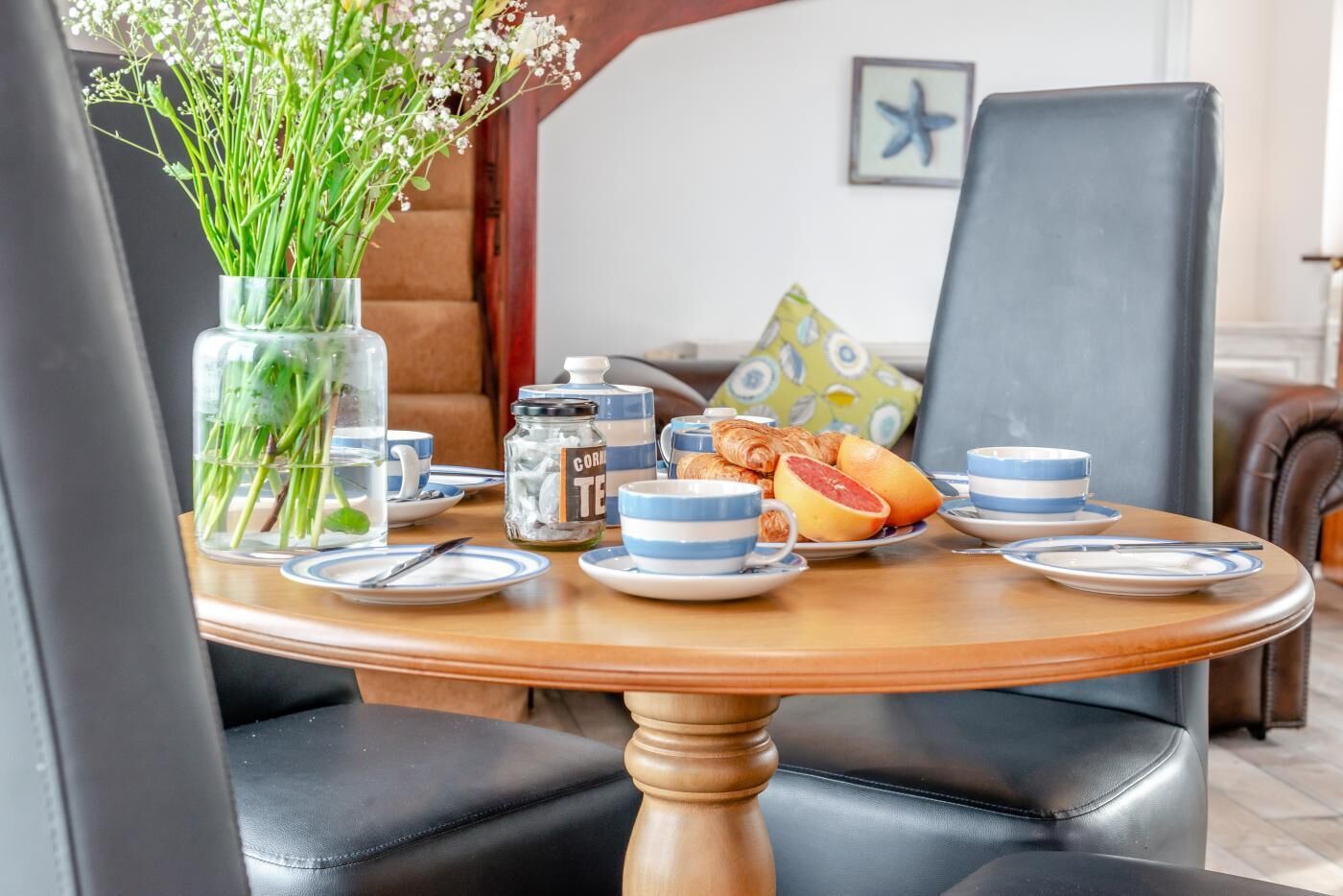 Une table ronde en bois, ornée d’un vase de fleurs, d’une cafetière, d’assiettes remplies de nourriture et de tasses.