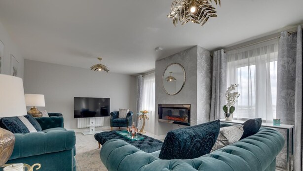 A modern living room with teal sofas, a flat-screen TV, a fireplace, and a large window with curtains. - 4 Bed in Seahouses (oc-gd1916) (Seahouses)