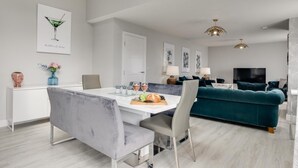 A modern living room with a teal sofa, a dining area with a white table, and a TV in the background. - 4 Bed in Seahouses (oc-gd1916) (Seahouses)