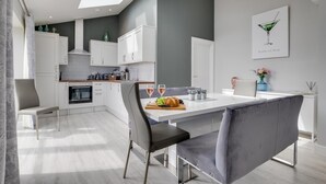 A modern kitchen with white cabinets, a dining table with grey upholstered chairs, and a large window above the sink. - 4 Bed in Seahouses (oc-gd1916) (Seahouses)