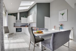 Une cuisine moderne avec des meubles blancs, une table à manger entourée de chaises à revêtement gris et une grande fenêtre au-dessus de l’évier.
