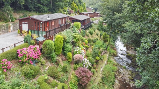 A hillside property with multiple wooden cabins, lush gardens, and a stream. - 2 Bed in Lynton (oc-r13823) (Barbrook)