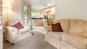 A cozy living room with two beige sofas and a coffee table, a kitchen in the background, and a window with curtains. - 2 Bed in Lynton (oc-r13823) (Barbrook)