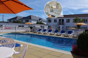 Ein Schwimmbecken mit Liegestühlen und Sonnenschirmen, ein weißes Gebäude mit Balkonen und ein eingezäunter Bereich.