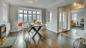 A spacious dining area with a wooden table, white chairs, and a view of the outdoors through large windows. - 4 Bed in Barmouth (oc-bow259) (Barmouth)