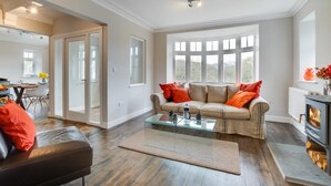 A modern living room with a glass coffee table, a beige sofa with orange cushions, and a fireplace. - 4 Bed in Barmouth (oc-bow259) (Barmouth)